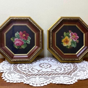 May include: Two framed floral cross-stitch artworks. Each piece features a colorful flower bouquet on a black background, set within an octagonal frame with a gold border and a dark wood-tone frame. The artworks are displayed on a wooden surface.