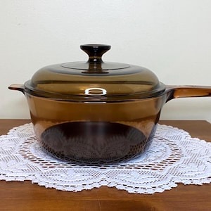 Può includere: Una casseruola vintage in vetro color ambra con coperchio e lungo manico. La pentola è realizzata in vetro trasparente, che consente la visibilità del contenuto. La casseruola è appoggiata su una tovaglietta di pizzo bianco.