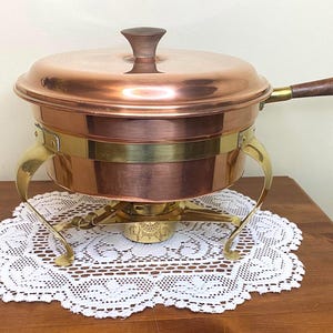 May include: A copper chafing dish with a brass stand and a wooden handle. The chafing dish is on a white lace doily.