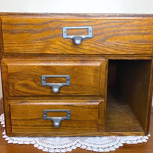 Puede incluir: Un mueble de almacenamiento de madera con tres cajones y un compartimento lateral. El mueble es de roble y tiene un acabado marrón dorado. Cada cajón tiene un portaetiquetas de metal. El mueble está sobre una mesa con un tapete de encaje blanco.