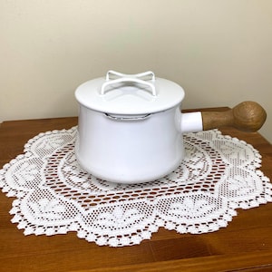 May include: A white enamel saucepan with a lid and a wooden handle. The saucepan is sitting on a white lace doily on a wooden table. The lid has a white handle.