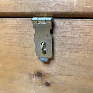 May include: Close-up of a wooden chest with a metal latch. The latch is a tarnished brass colour and is attached to the chest with a hinge. The wood grain is visible, and the chest appears to be made of pine.