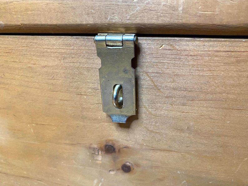 May include: Close-up of a wooden chest with a metal latch. The latch is a tarnished brass colour and is attached to the chest with a hinge. The wood grain is visible, and the chest appears to be made of pine.