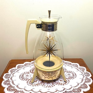 May include: A vintage glass coffee pot with a gold star design and a matching cream-coloured warming base. The pot has a clear glass body and a cream-coloured handle.