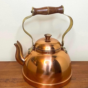 Peut inclure: Une bouilloire en cuivre avec une poignée en laiton et un bouton en bois. La bouilloire a une finition brillante et polie.