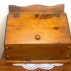 Könnte beinhalten: Ein Schreibtisch-Organizer aus Holz mit einem Klappdeckel und einem kleinen Holzknopf. Der Organizer ist aus hellbraunem Holz gefertigt und hat Messing Scharniere. Die Oberseite hat ein geschwungenes Design. Eine weiße Spitzendecke ist unten sichtbar.