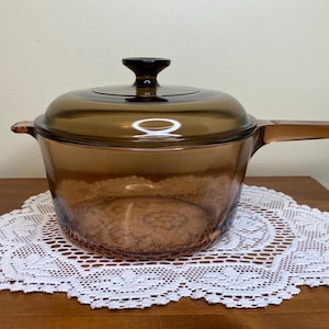 Può includere: Una casseruola vintage in vetro color ambra con coperchio e lungo manico. La casseruola è appoggiata su una tovaglietta di pizzo bianco su una superficie di legno. Il coperchio ha un pomello nero.