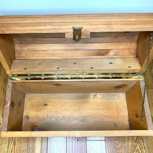 May include: Open wooden chest with a hinged lid and brass fittings. The chest is made of light-coloured wood and has a rectangular shape. A small chain is attached to the lid to prevent it from opening too far. The interior is empty.