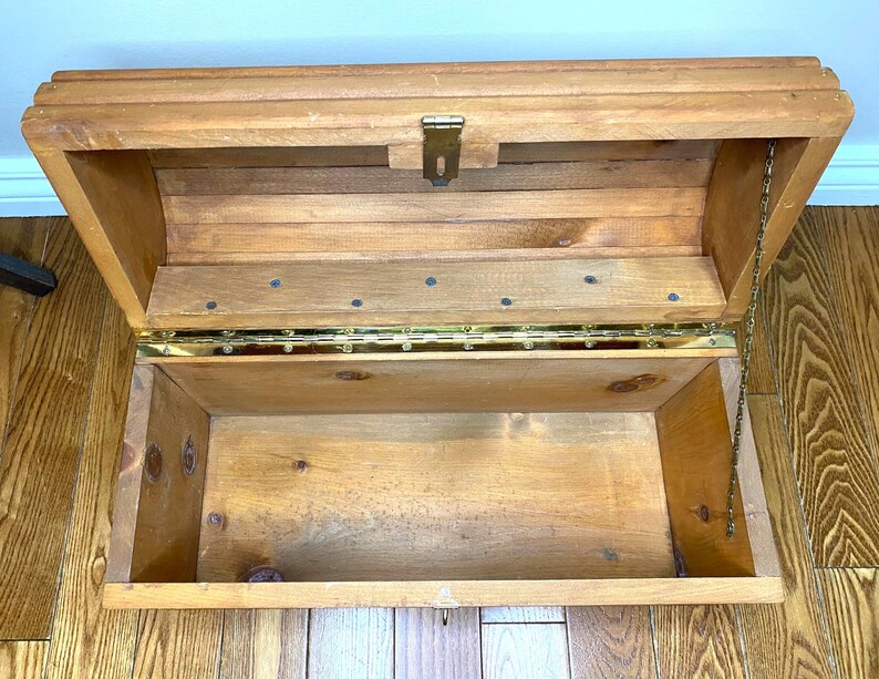 May include: Open wooden chest with a hinged lid and brass fittings. The chest is made of light-coloured wood and has a rectangular shape. A small chain is attached to the lid to prevent it from opening too far. The interior is empty.