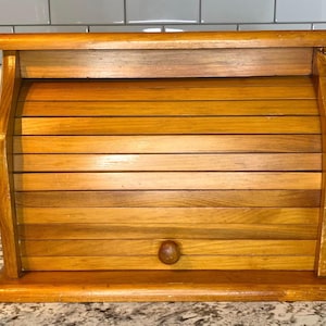 Può includere: Una cassetta per il pane in legno con porta a rullo. La cassetta è realizzata in legno di colore chiaro e ha un frontale arrotondato. Un piccolo pomello rotondo in legno è sul davanti. La cassetta per il pane è progettata per mantenere il pane fresco.