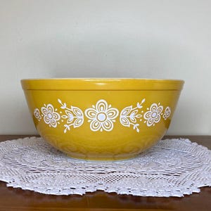 Op de afbeelding: Een mosterdgele mengkom met een herhalend wit bloemen- en bladpatroon rond de buitenkant. De kom heeft een gladde, afgeronde rand en een brede, diepe kom, wat een klassiek ontwerp met een levendige, effen kleurafwerking laat zien.