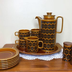 May include: A ceramic coffee set featuring a tall coffee pot, a creamer, two cups, and two matching canisters, all decorated with a dark brown and amber geometric pattern. A stack of six plain brown saucers sits beside the tray.