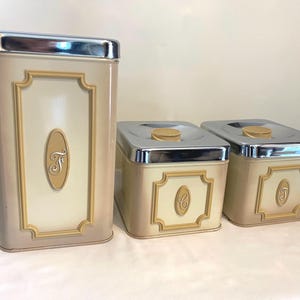 Può includere: Set di tre barattoli da cucina vintage con coperchi cromati. I barattoli sono di colore crema con un bordo beige e hanno una lettera 'F' sul davanti di ciascuno.