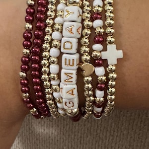 May include: A stack of beaded bracelets in shades of burgundy, gold, and white. One bracelet spells out "GAMEDAY" with white letter beads. Other bracelets feature gold beads, burgundy beads, and a white cross charm.