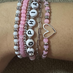 May include: A set of six bracelets, including a silver chain bracelet with a heart charm, and five beaded bracelets. The beaded bracelets are made with white and pink beads, and one of the bracelets has white beads with the name "SOPHIA" spelled out in black letters.