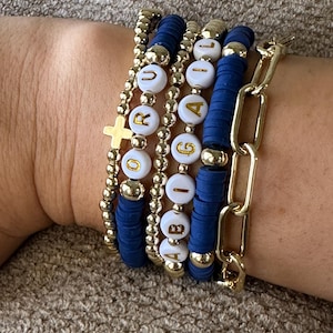 May include: A set of five bracelets featuring gold beads and white beads with gold lettering spelling out the name "Abigail". The bracelets are stacked on a person's wrist and include a gold chain bracelet.