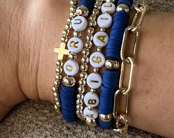 Op de afbeelding: Een set van vijf armbanden met gouden kralen en witte kralen met gouden letters die de naam "Abigail" spellen. De armbanden zijn gestapeld op de pols van een persoon en bevatten een gouden kettingarmband.