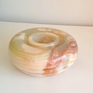 Könnte beinhalten: Ein runder Kerzenhalter aus Naturstein mit einer zentralen Vertiefung. Der Stein weist Schichten in Creme-, Beige- und Rotbrauntönen auf. Ein dekorativer Artikel für die Inneneinrichtung.