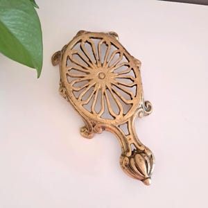 Peut inclure: Un dessous de plat doré orné d'un motif ajouré décoratif. Le dessous de plat est doté d'une poignée et d'un motif circulaire avec des lignes rayonnantes. La pièce métallique est probablement en laiton ou en alliage similaire.