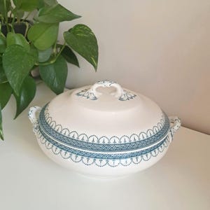 May include: A white ceramic serving dish with a lid, featuring a blue decorative border and handles. The dish has a rounded shape and is placed on a white surface. The lid has a handle and a matching blue design.