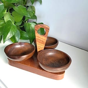 May include: A dark brown wooden serving tray featuring three circular bowls attached to a central vertical handle with a circular cutout. The wood grain is visible throughout the structure, which is composed of a flat base and three bowls.