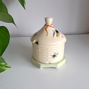 May include: A ceramic honey pot shaped like a beehive, with a cream-colored body and a light green base. The lid is adorned with orange and brown accents and painted bees. The pot has a small opening for a spoon.