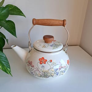 Op de afbeelding: Witte emaille theepot met een bloemenontwerp met rode, paarse en blauwe bloemen. Het heeft een houten handvat en dekselknop, en een zilveren metalen handvat. Een klassiek keukenartikel.
