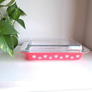 Könnte beinhalten: Ein rechteckiges, rotes Pyrex-Backgeschirr mit einem Deckel aus klarem Glas. Das Geschirr ist mit weißen Schneeflockenmustern an den Seiten verziert. Das Geschirr steht auf einer weißen Oberfläche.
