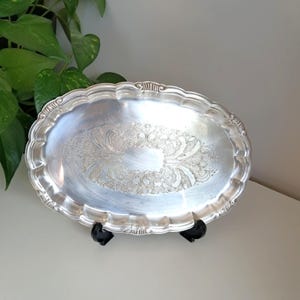 Può includere: Un vassoio ovale in metallo color argento con bordo smerlato e un intricato motivo floreale in rilievo al centro. Il vassoio è sostenuto da un supporto in plastica nera e presenta una superficie metallica riflettente con volute dettagliate.