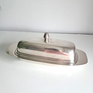 Può includere: Un portaburro in metallo color argento lucido con una base ovale allungata e un coperchio a cupola abbinato. Il coperchio presenta un piccolo pomello arrotondato al centro, che funge da maniglia per sollevare la copertura dalla base.