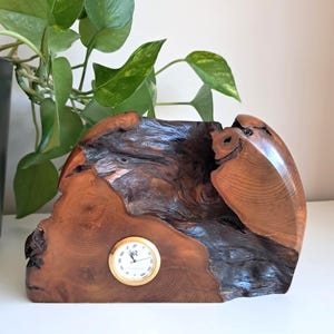 May include: A wooden clock with a natural, organic form. The clock has a round, gold-coloured clock face with black numerals and hands. The wood displays rich brown and dark tones, highlighting the natural grain and texture. A decorative item for a desk or shelf, made from natural wood.