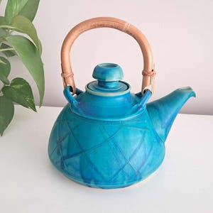 Puede incluir: Una tetera de cerámica azul turquesa con un asa de bambú tejido y una tapa. La tetera tiene un patrón geométrico grabado en la superficie.