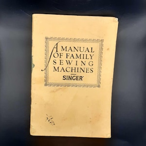 Könnte beinhalten: Ein vergilbtes Vintage-Handbuch für Singer-Nähmaschinen für die Familie. Der Umschlag zeigt einen dekorativen Rahmen und den Text "A Manual of Family Sewing Machines - Singer".