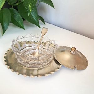 Peut inclure: Un bol en verre pressé transparent avec un bord festonné repose sur un plateau rond en métal argenté au bord festonné assorti. Une petite cuillère en métal argenté est placée dans le bol, accompagnée d'un couvercle rond en métal argenté avec un bouton.