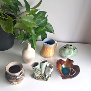 May include: A selection of handcrafted ceramic vases and decorative items in diverse forms and hues. Features a small jug, a spherical vase with apertures, and a heart-shaped dish. A potted plant is visible in the background.