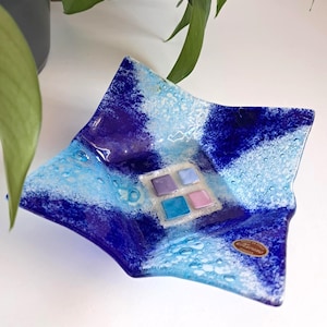 May include: A square glass dish with a textured surface and a vibrant color scheme. The dish features shades of blue and white, with a central square containing four smaller squares in purple, blue, and pink. A decorative piece for the home.