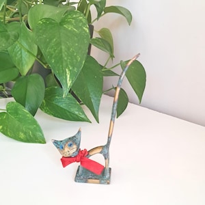 Op de afbeelding: Een kleine bronzen kattenfiguur met een rode sjaal. De kat heeft blauwe ogen en een lange, gebogen staart. Het beeldje staat op een wit oppervlak met groene planten op de achtergrond.