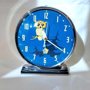 Puede incluir: Un reloj despertador vintage de cromo con una esfera azul y una ilustración de búho. La esfera del reloj tiene marcadores de hora en números romanos negros y marcadores de minutos en números arábigos negros. El búho es blanco con ojos amarillos y está posado en una rama con una pequeña casa en el fondo.