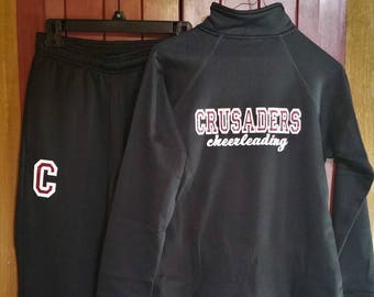 以下が含まれることがあります： 黒いスウェットパンツと黒いジップアップスウェットシャツ。背面に「Crusaders cheerleading」という文字が白と赤で書かれています。