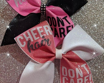 May include: Two cheer bows with glitter fabric. The top bow is black and pink with the text "Cheer hair don't care". The bottom bow is white and pink with the text "Cheer hair don't care!"