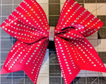 May include: A vibrant red cheerleading bow adorned with numerous sparkling rhinestones. The bow features a central rhinestone band and is designed with multiple layers of red fabric. The rhinestones are arranged in rows and clusters, adding a glamorous touch.