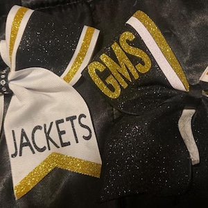 May include: Two cheerleading bows. One is white with black and gold accents, the word "Emory" and "JACKETS" in black. The other is black with gold and white accents, with "GMS" and "JACKETS" in gold.