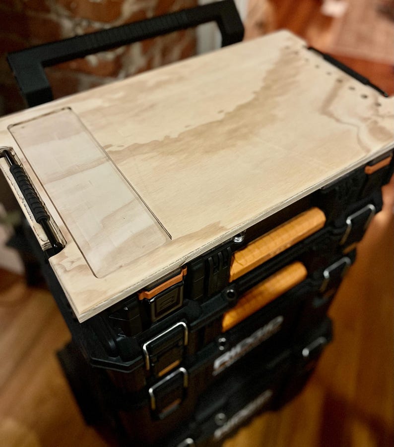 Op de afbeelding: Een zwarte gereedschapskist met een houten bovenkant. De bovenkant heeft een rechthoekige uitsparing en is vastgeschroefd. De kist heeft meerdere laden en staat op wielen.