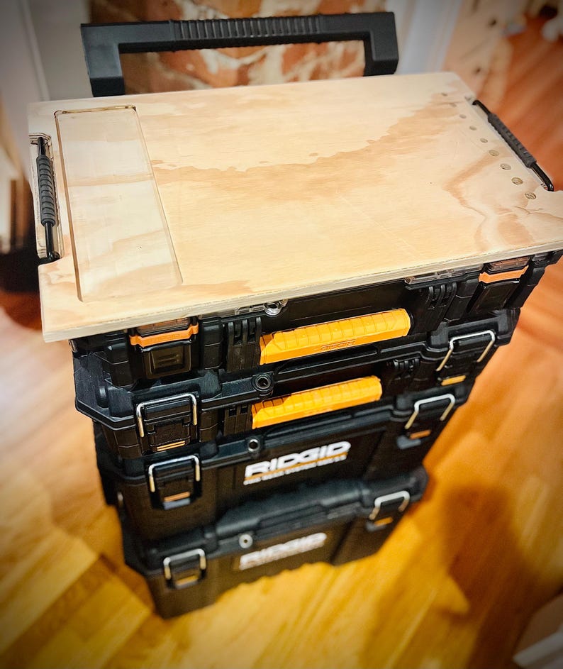 Op de afbeelding: Een stapel van drie zwarte gereedschapskisten met oranje handvatten en het woord "RIDGID" erop gedrukt. De bovenste gereedschapskist heeft een houten bovenkant met een uitsparing voor een gereedschap.