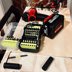 Op de afbeelding: Een rode en zwarte accuboormachine met een accu en een koffer met boorbits. De boormachine ligt op een houten oppervlak met een bakstenen muur op de achtergrond.