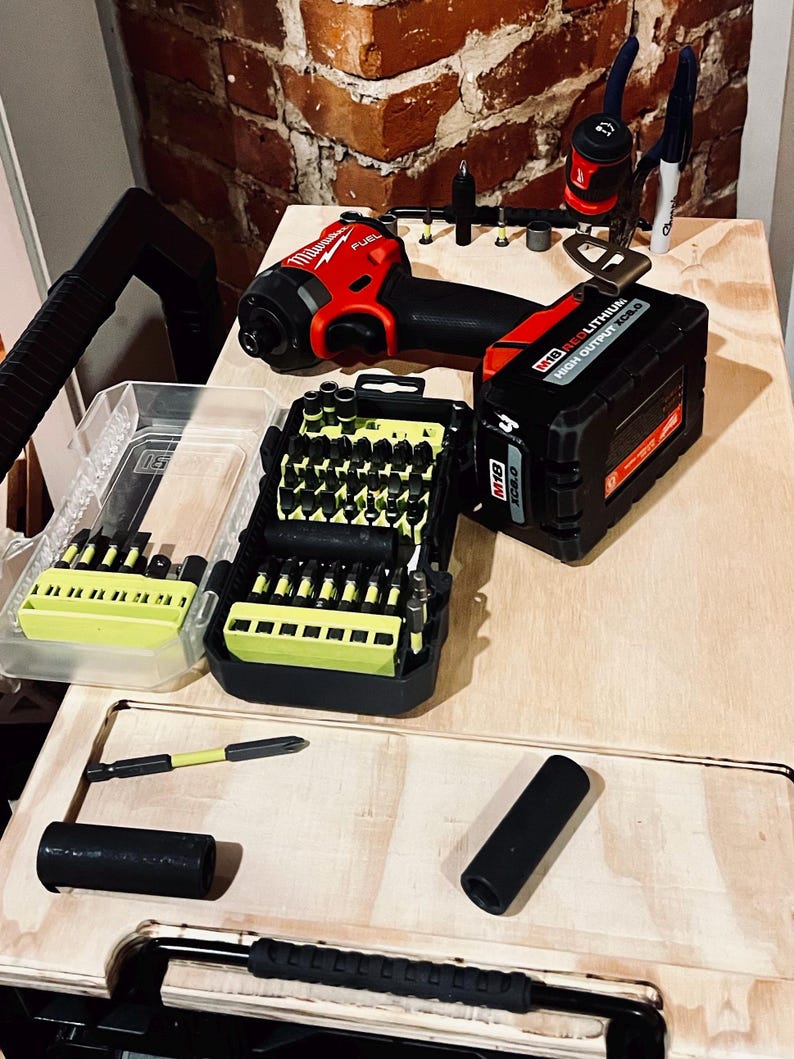 Op de afbeelding: Een rode en zwarte accuboormachine met een accu en een koffer met boorbits. De boormachine ligt op een houten oppervlak met een bakstenen muur op de achtergrond.