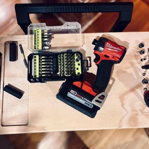 Op de afbeelding: Een rode en zwarte draadloze boormachine met een Milwaukee FUEL-label, een rode lithium-accu en een koffer met verschillende boorbits.