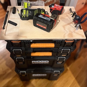 Op de afbeelding: Een zwarte en oranje gereedschapskist met een houten bovenkant. De bovenkant heeft een Milwaukee-boormachine, een batterij en een verscheidenheid aan gereedschap.