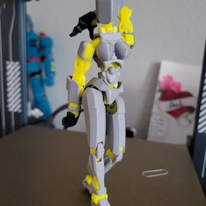 Puede incluir: Una figura de acción articulada impresa en 3D con un esquema de colores gris y amarillo. La figura tiene una cabeza lisa sin rostro, un torso segmentado y extremidades articuladas. El diseño incluye placas grises sobre un marco amarillo.