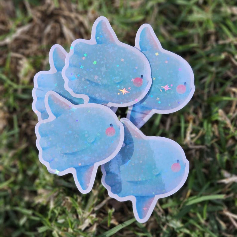 Peut inclure: Quatre autocollants bleu et blanc iris&eacute;s repr&eacute;sentant un poisson-lune de dessin anim&eacute; avec une texture scintillante. Les autocollants sont dispos&eacute;s en grappe sur une surface herbeuse verte.